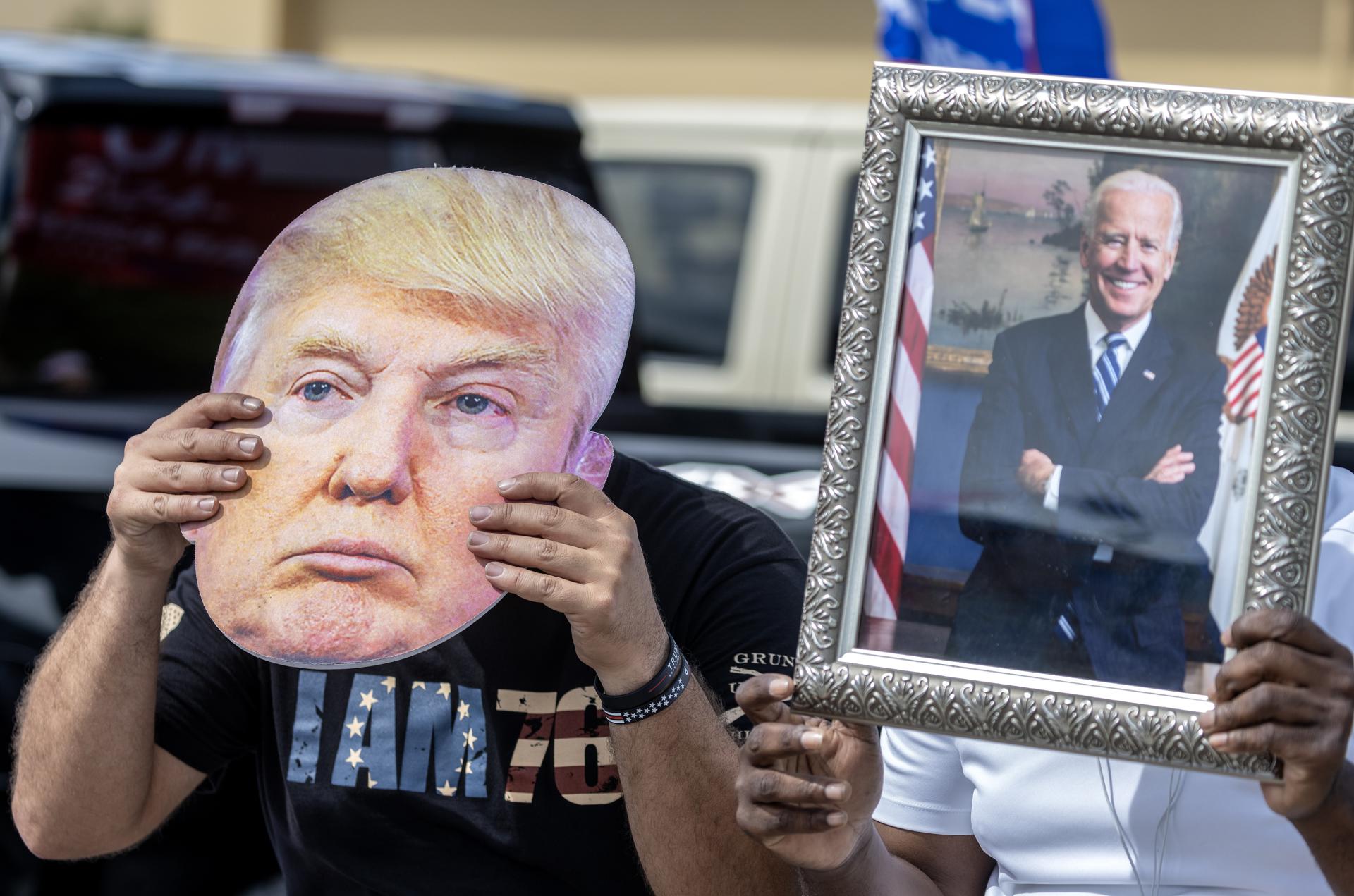 CAMPAÑA. Un partidario de Joe Biden sostiene un retrato del presidente estadounidense (der) junto a un seguidor de Donald Trump que lleva una foto del exmandatario. EFE